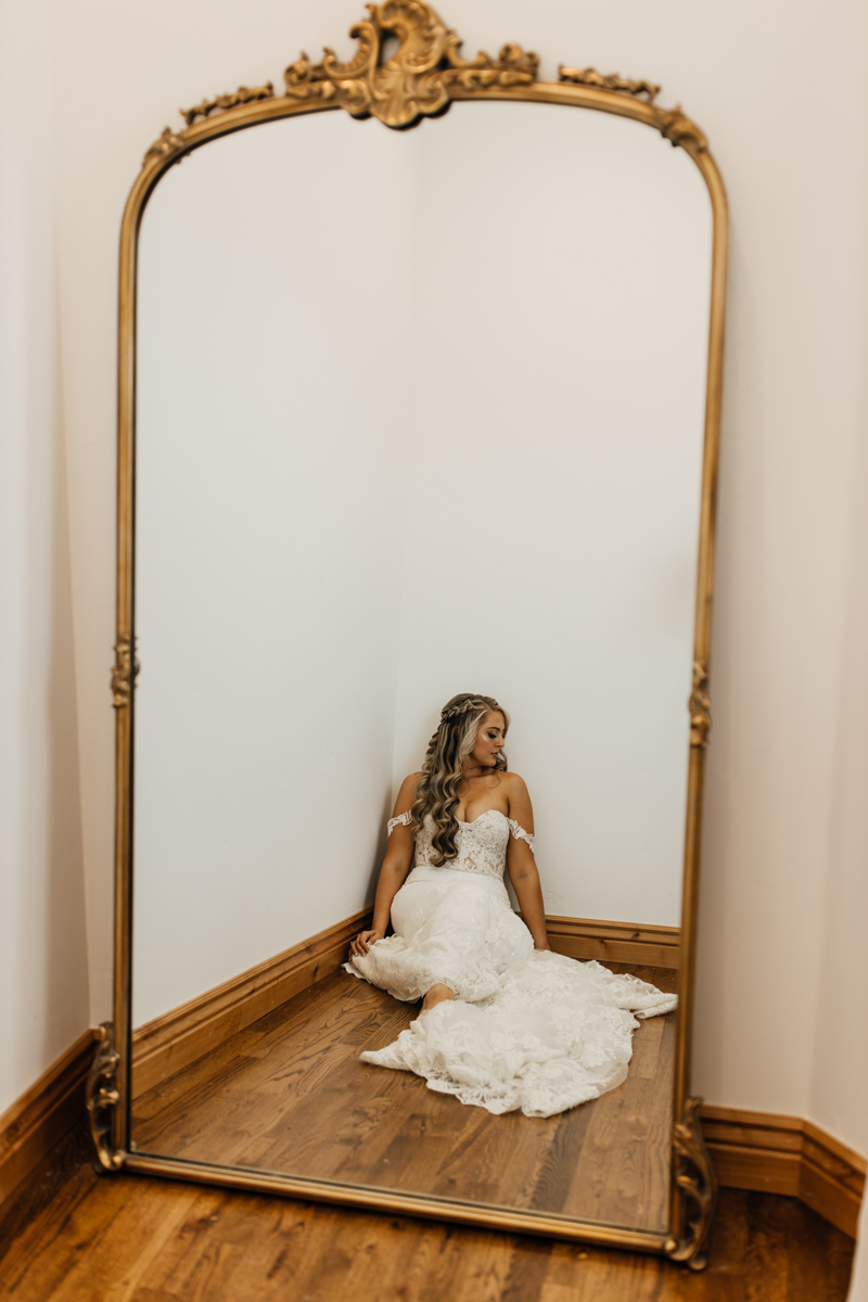 Photography Studio on Main, a woman is reflected in a mirror with gold frame, she sits in the corner of the room contemplating in a dress
