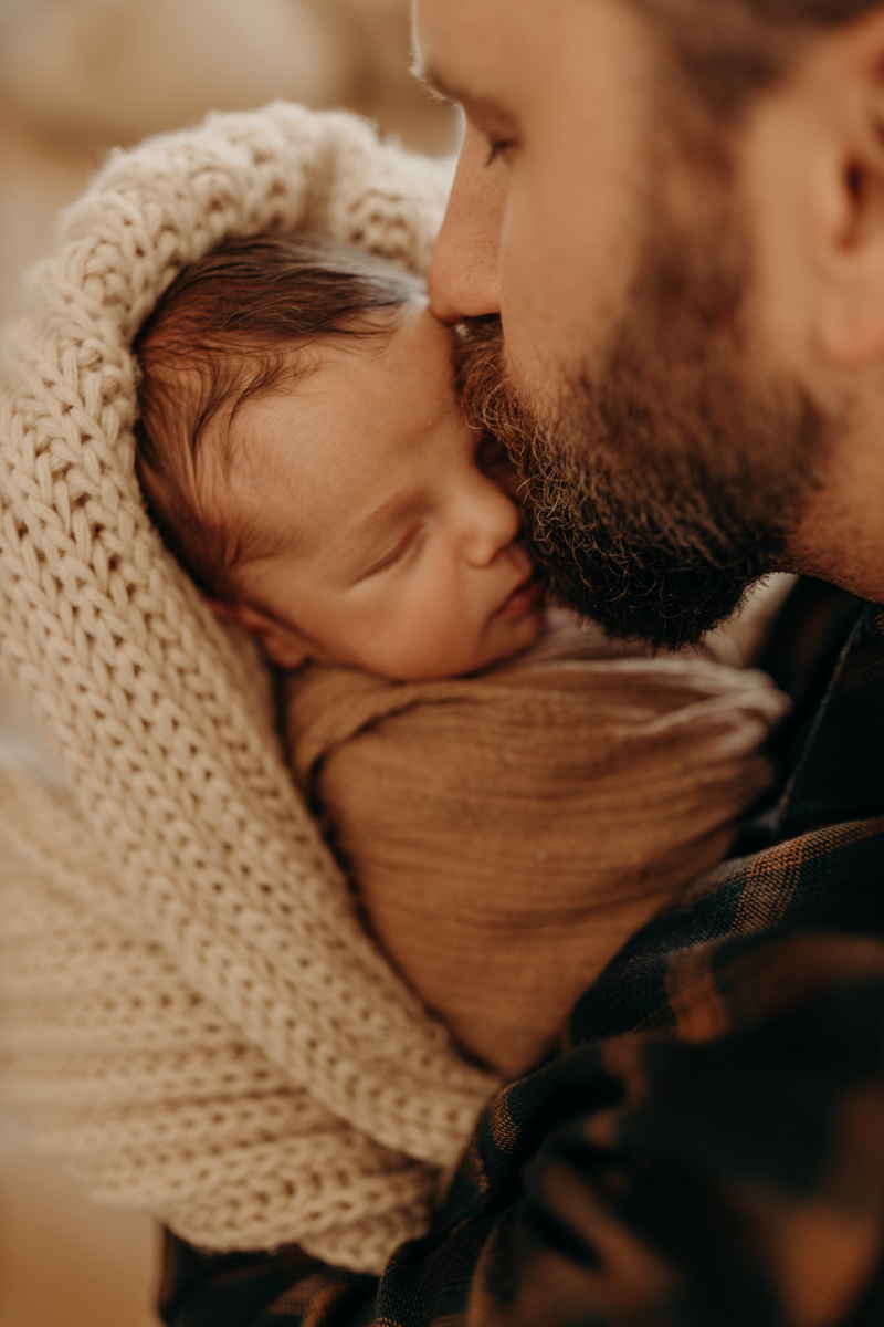 Newborn Photography, a father kisses his young baby swaddled in his arms