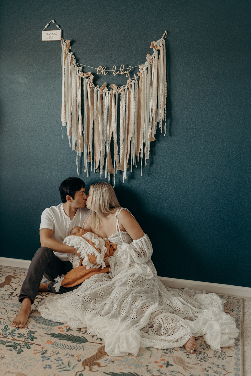 Newborn Photography, a man and woman kiss as they sit on the floor and hold baby