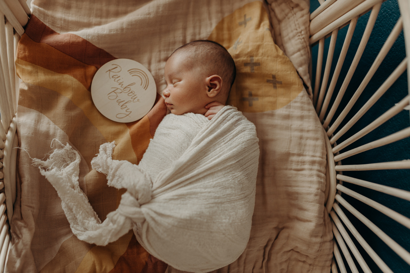 Newborn Photography, a little baby sleeps atop blankets in a wicker basket, with a sign beside that says "Rainbow Baby"