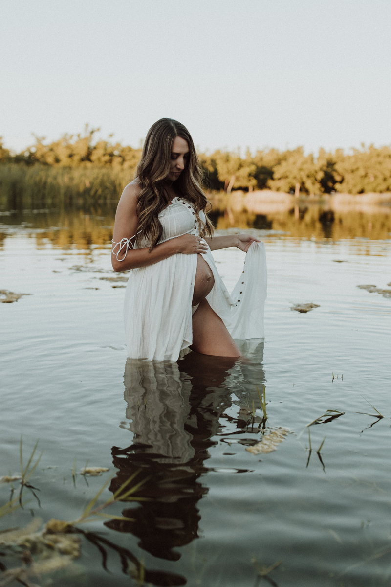 Maternity Photography, an expecting woman stands knee deep in a lake while holding up her dress to walk
