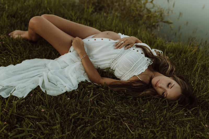 Maternity Photography, a woman lays in the grass the near lake in a dress, she is pregnant