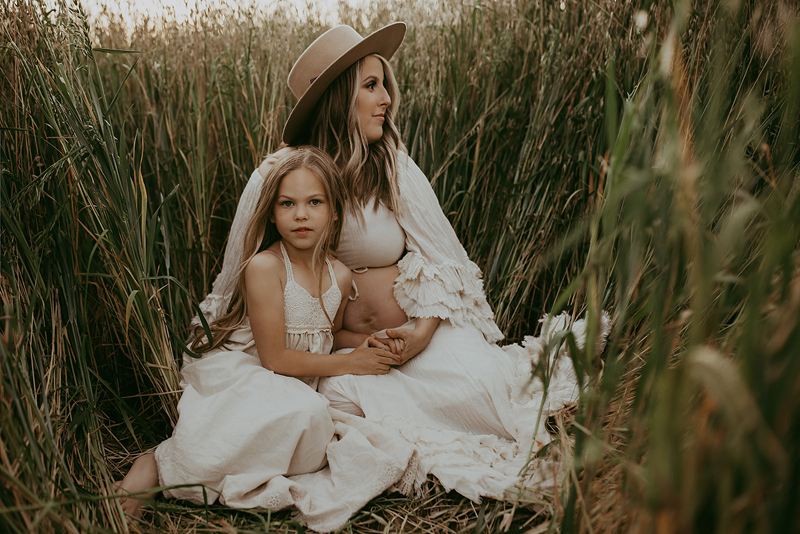 Family Photography, a mother sits among flattened reeds, her daughter beside her, they are both in white dresses