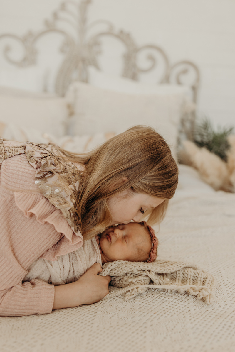 Newborn Photography,  a young girl kisses her new baby sister who sleeps comfortable atop a freshly made bed