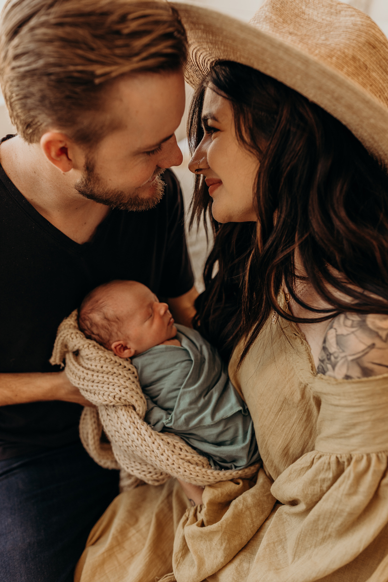 Newborn Photography, a young mom and dad smile at each other face to face as mom holds her new baby