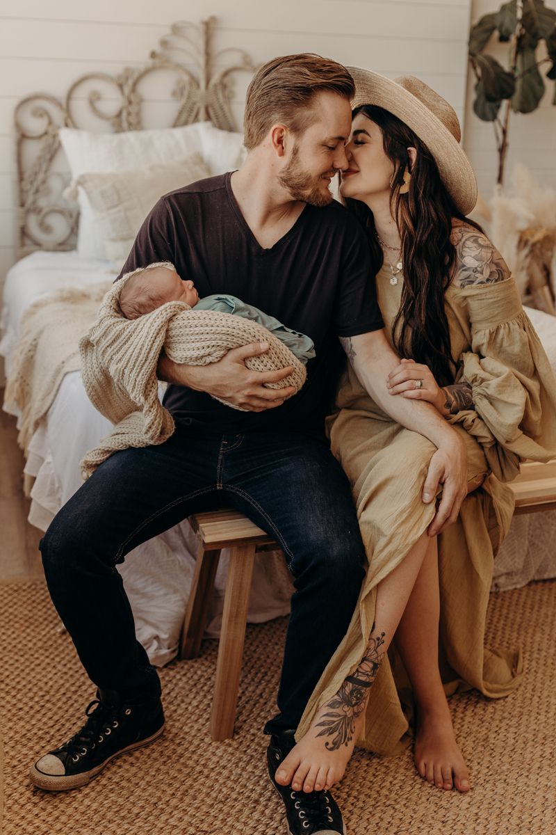 Newborn Photography, a man and woman lovingly lean into each other at the foot of a bed, dad holds baby