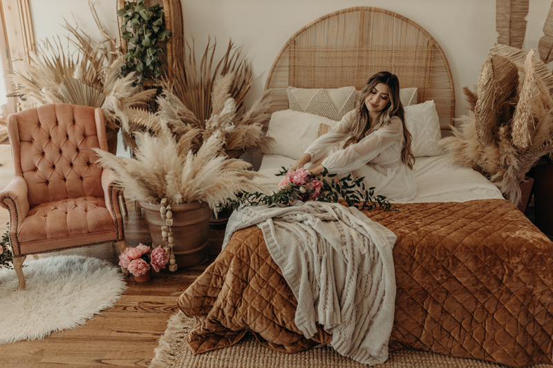 Photography Studio on Main, a woman sits on a bed with flowers beside her content, the room is decorated with pampas grass and boho aesthetics