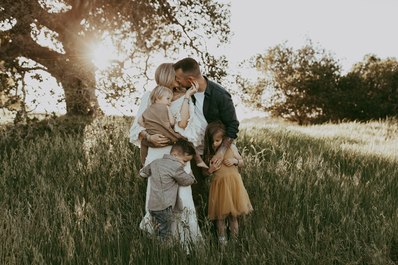 Family Photography, a husband kisses his wife and holds his children in a grassy meadow