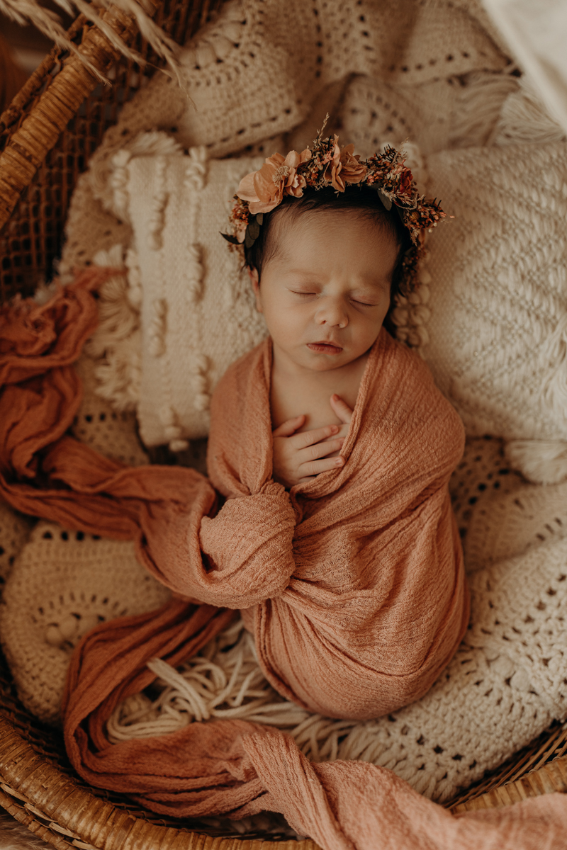 Newborn Photography,  a young baby girl is wrapped blankets and lay cozily in a wicker basket, floral crown atop her head