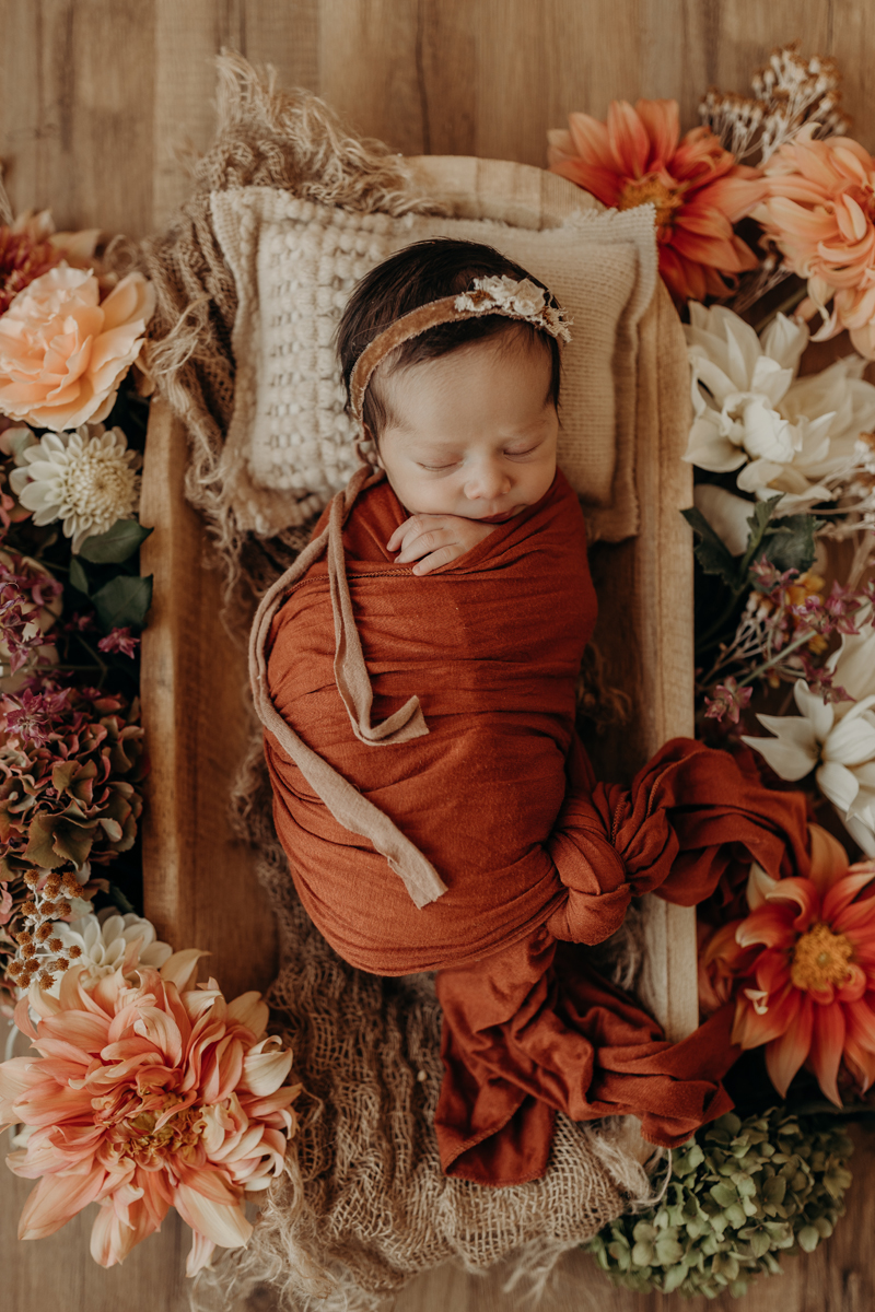 Newborn Photography, a lttle baby is wrapped in linens and surrounded by flowers in a small wicker basket
