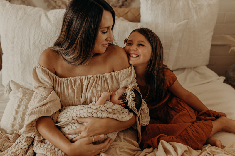 Newborn Photography, a mother sits in her bed holding her new daughter, older sister leans in smiling