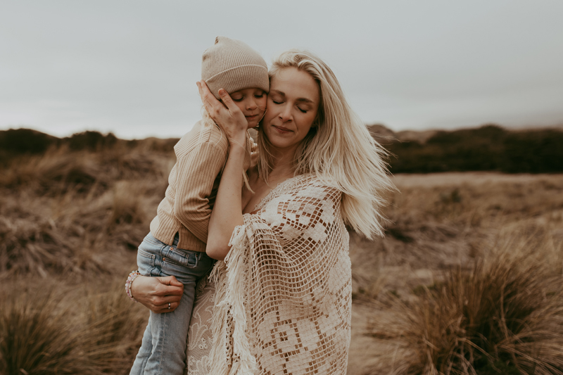 Family Photography, a mother holds her daughter cheek to cheek near a grassy beachside