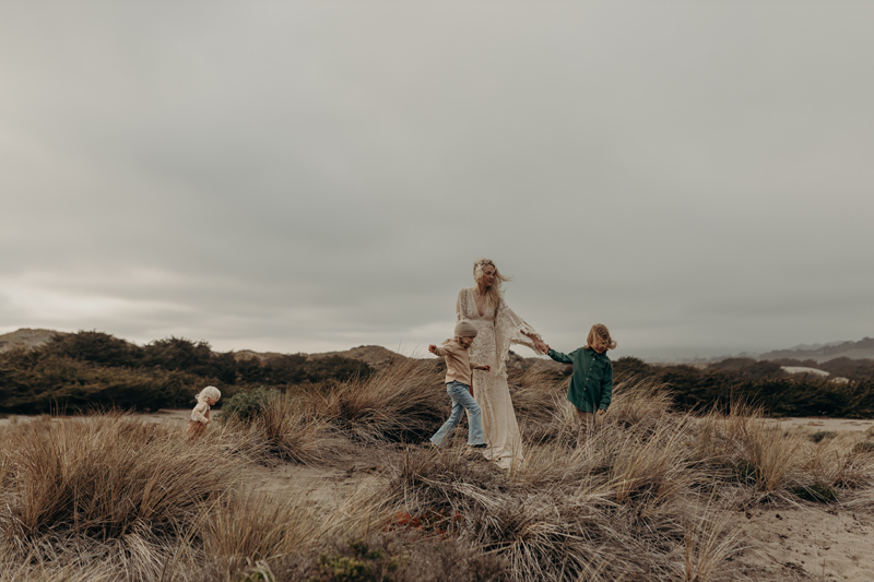 Family Photography,  a young mother in a dress walks through a grassy beach with her three children