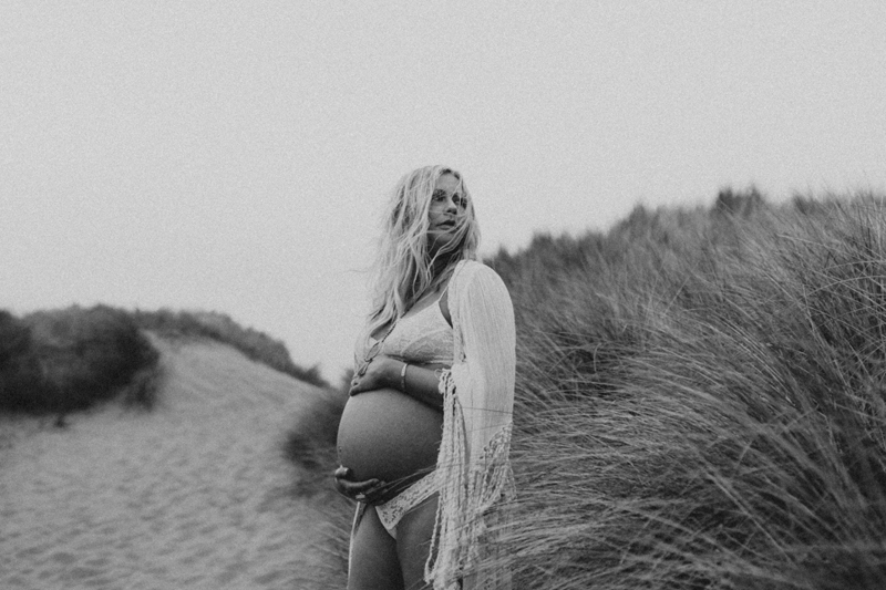 Maternity Photography, a woman stands on a hilly beach among tall grass, she holds her belly in final trimester