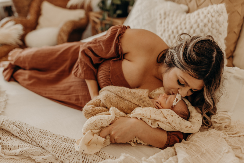Newborn Photography, a woman lays on a bed and kisses her new baby daughter sleeping in her arms