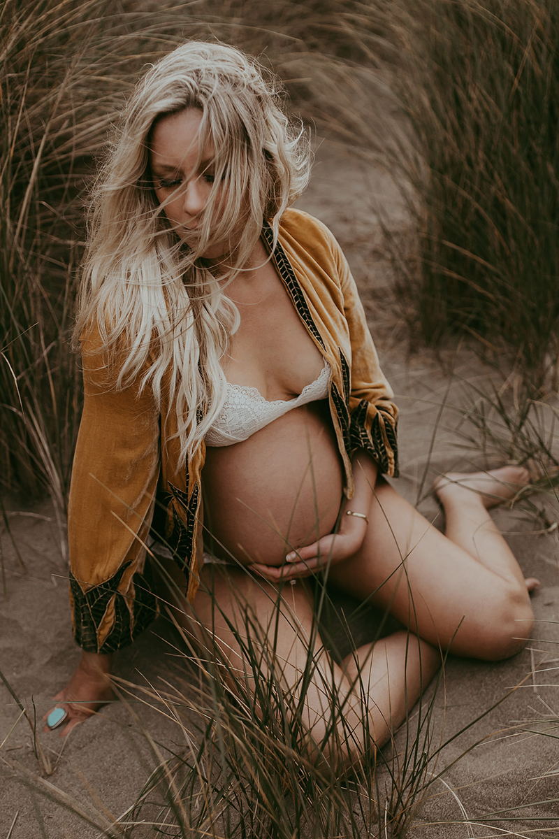 Maternity Photography, a pregnant woman sits in the sand near lake reeds holding her belly