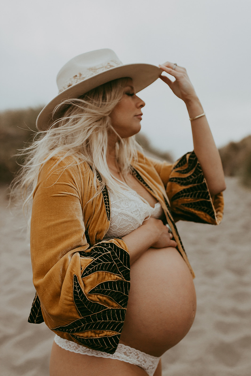Maternity Photography, a woman stands on a sandy beach while holding her belly, she is pregnant