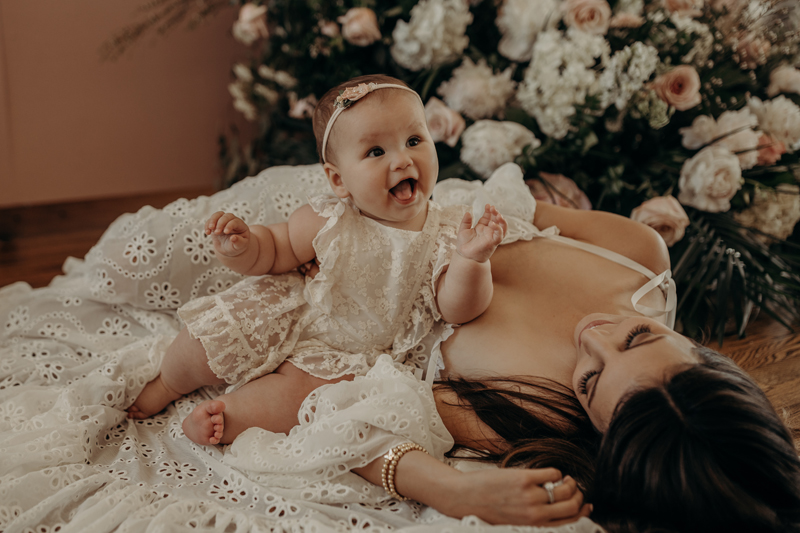 Family Photography, a mother in a dress lays on the floor beside a floral arrangement with baby sitting beside her smiling